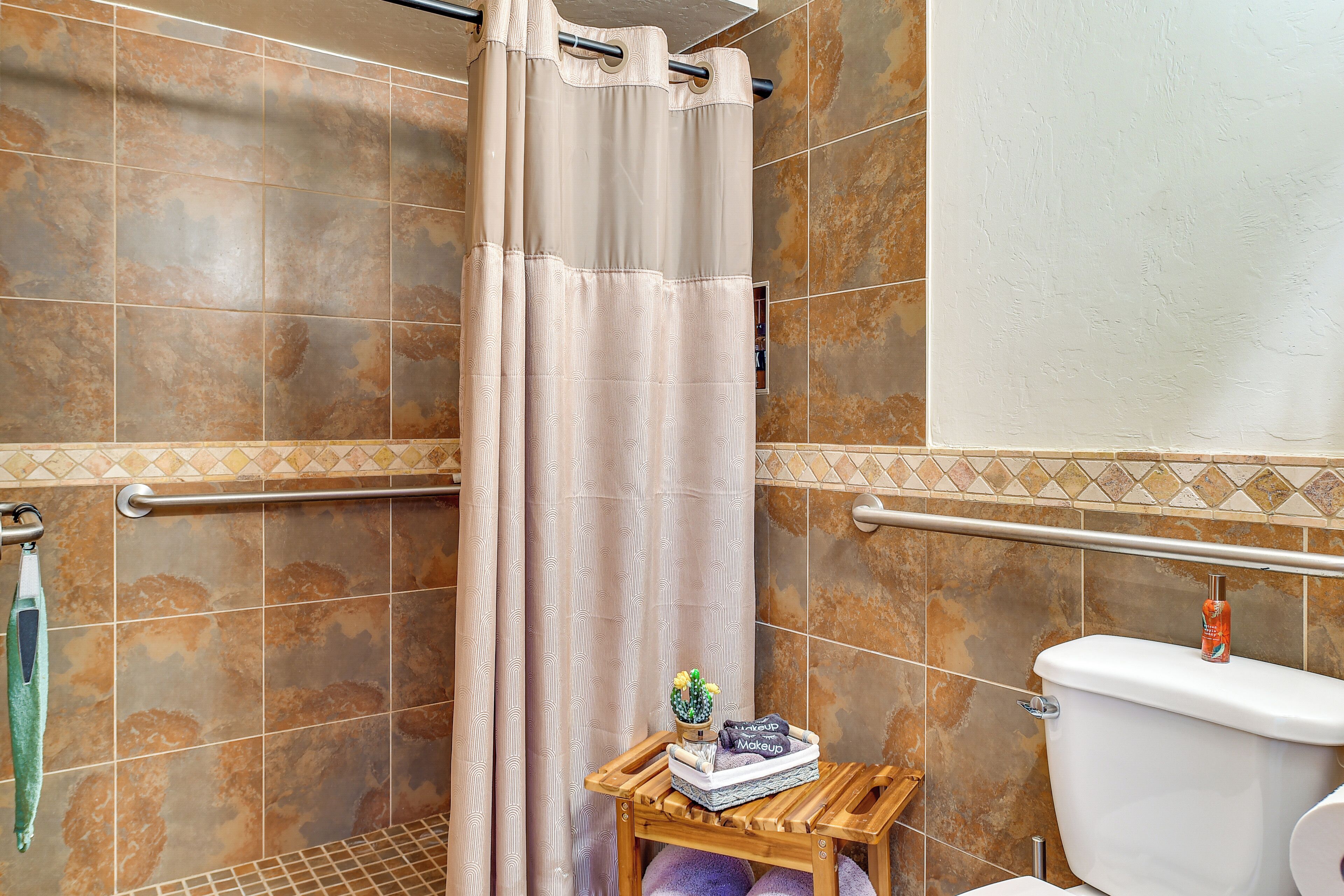 Une salle de bain avec un rideau de douche, des toilettes, une petite table sur laquelle se trouvent un cactus et des produits de maquillage, et une barre d’appui.
