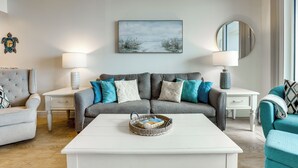 Ein Wohnzimmer mit weißem Couchtisch, grauem Sofa mit Türkis-Kissen, einem weißen Beistelltisch mit Lampe und einem gerahmten Kunstwerk an der Wand.