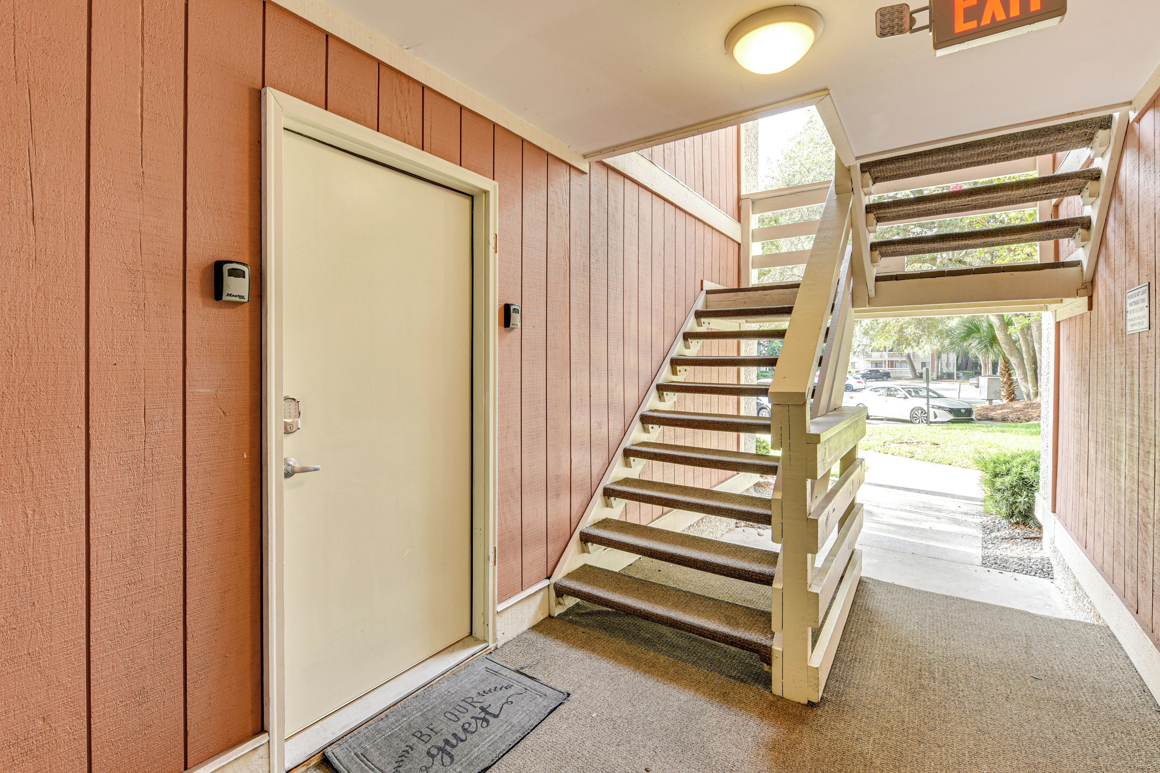 A wooden staircase leading to an upper level, a door with a keypad lock, and a doormat with a message.