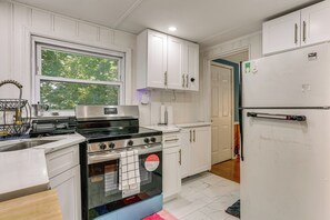 Een keuken met witte kasten, een roestvrijstalen fornuis, een koelkast en een raam met uitzicht op bomen.