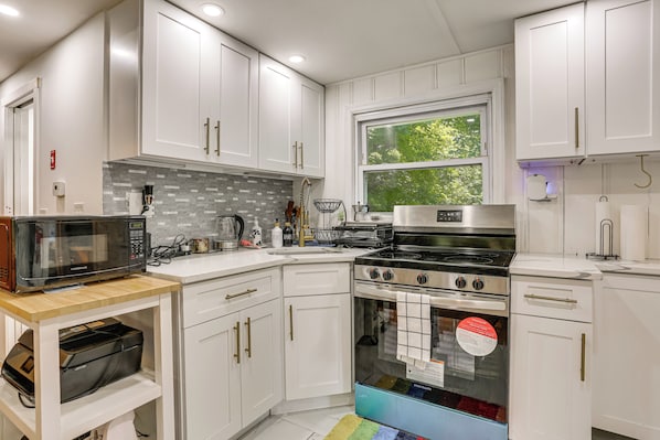A modern kitchen with white cabinets, a stainless steel stove, a microwave, and a window with a view of trees.