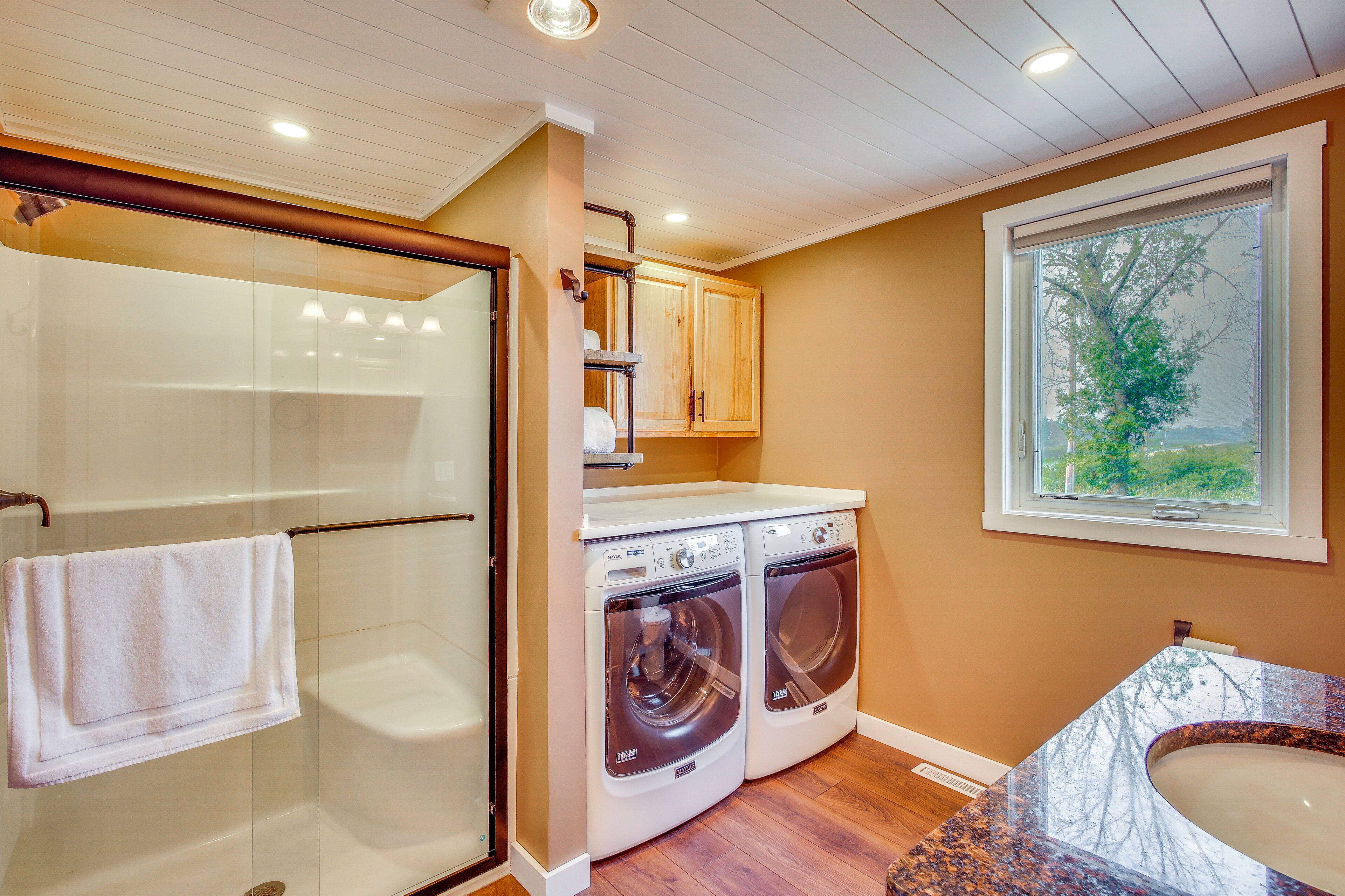 A laundry room with two front-loading washing machines, a glass shower enclosure, and a window with a view of trees.