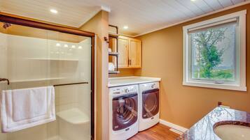 A laundry room with two front-loading washing machines, a glass shower enclosure, and a window with a view of trees.