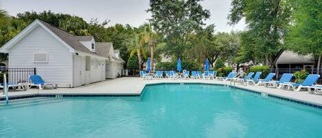 A swimming pool with blue lounge chairs and umbrellas, surrounded by trees and a white building.