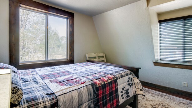 Une chambre avec une grande fenêtre, un lit recouvert d’une couvre-lit à motifs, une tête de lit en bois et un ventilateur de plafond.