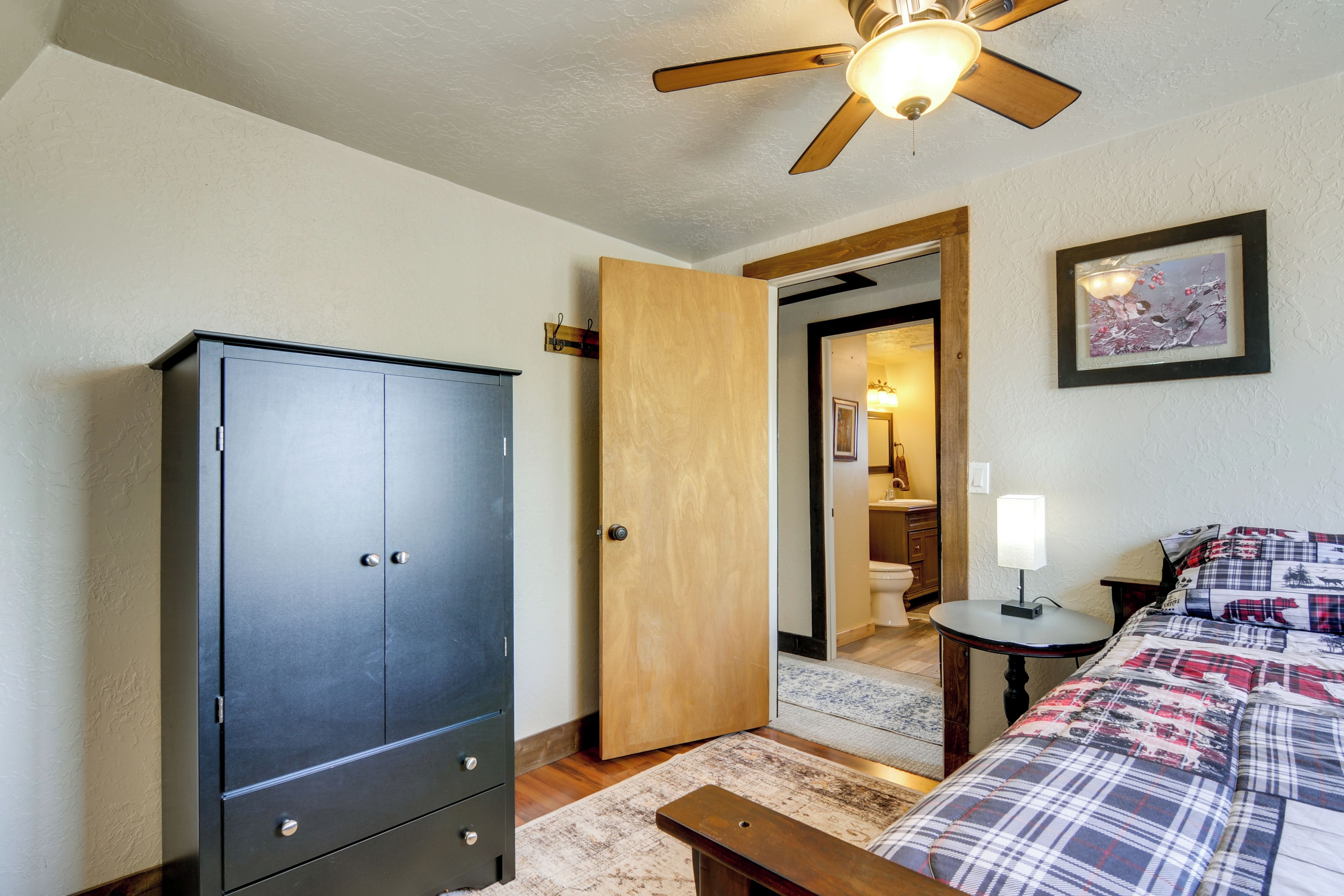 Une chambre à coucher avec une armoire, un lit recouvert d’une couette à carreaux, une table de chevet avec une lampe et un ventilateur de plafond.