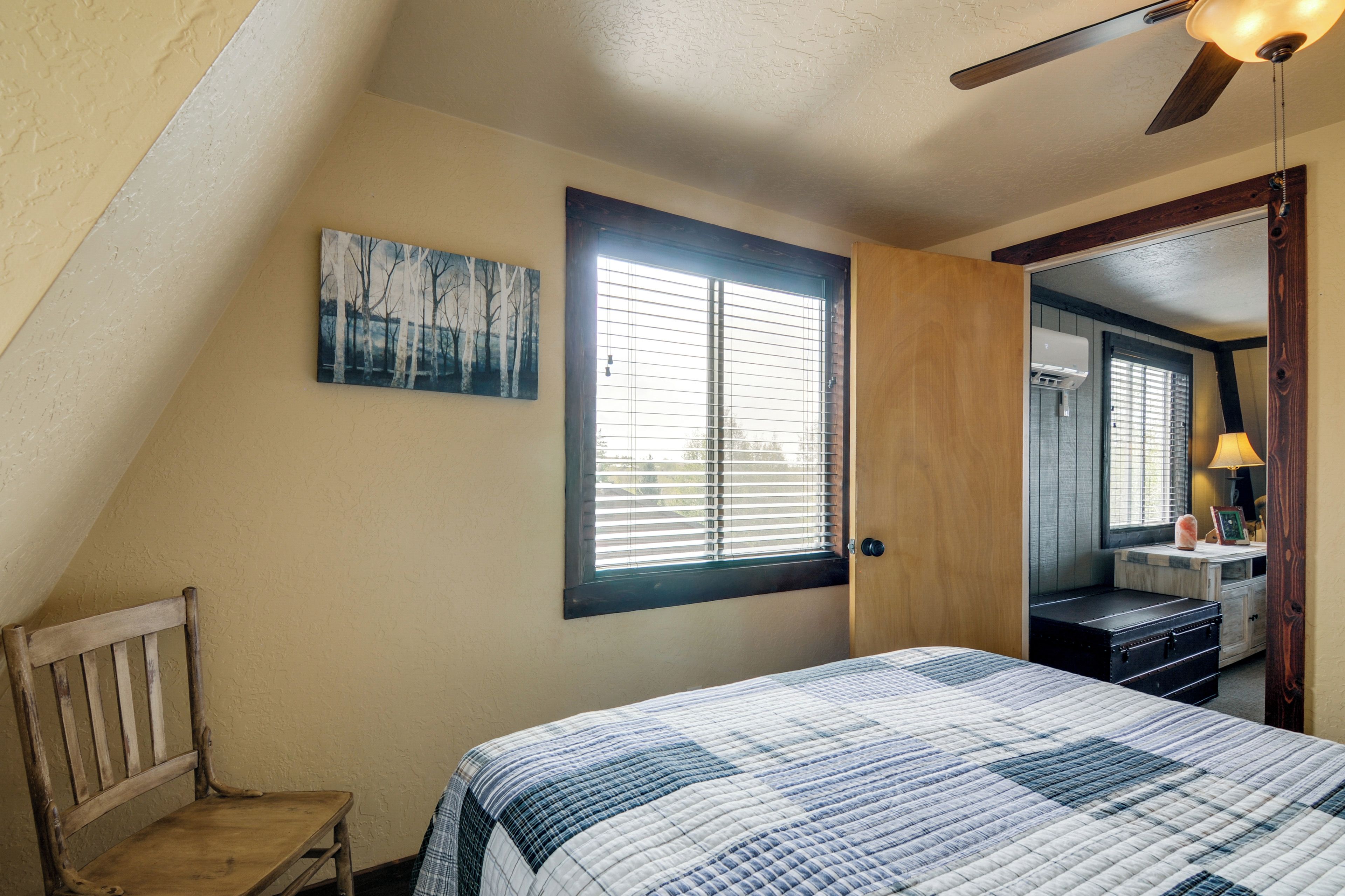Un dormitorio con techo inclinado, una cama con colcha, una silla de madera, un ventanal con persianas y una puerta que da a otra habitación.
