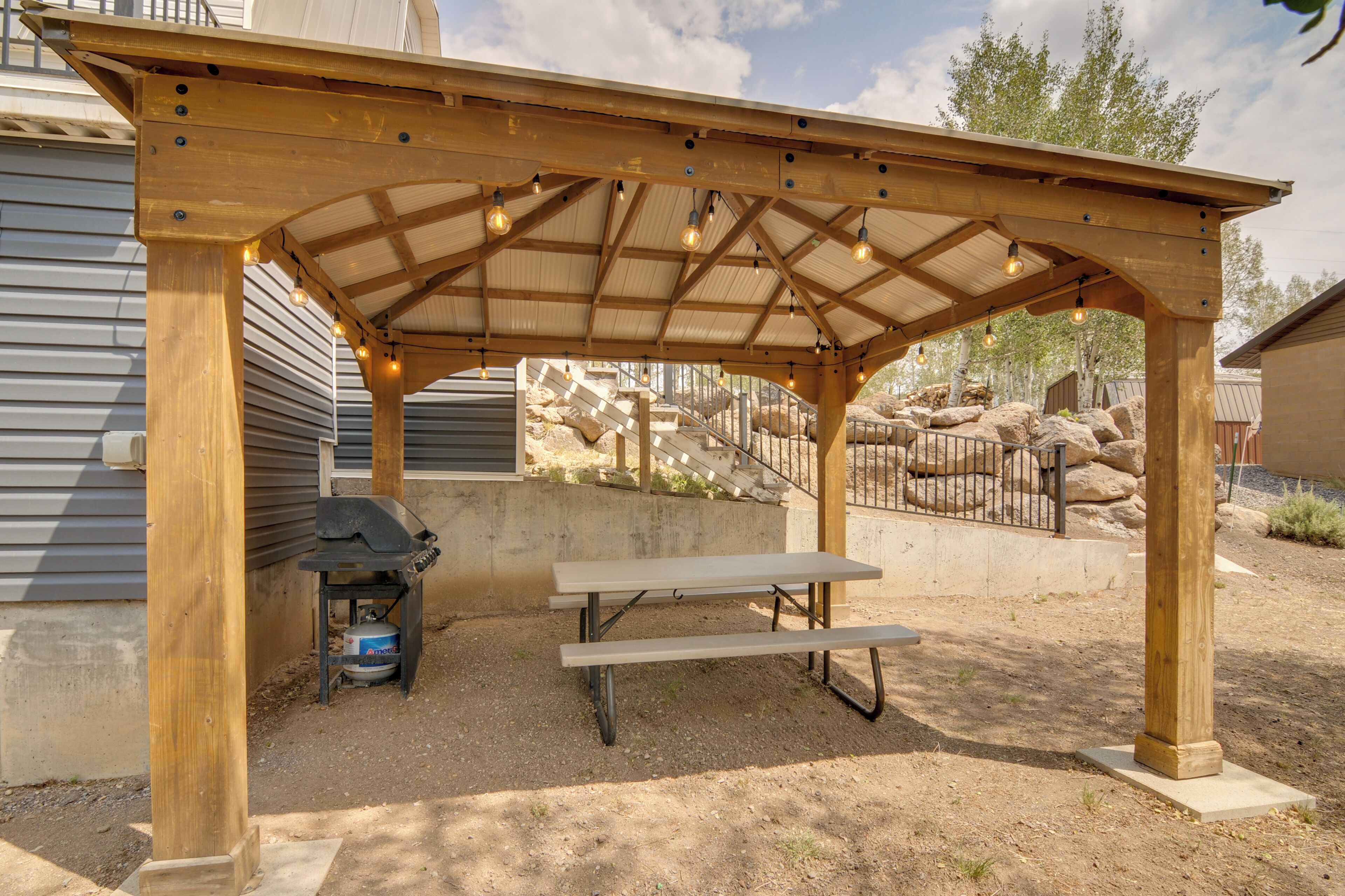 Una pérgola de madera con luces colgantes, una parrilla y una mesa de picnic en un jardín.
