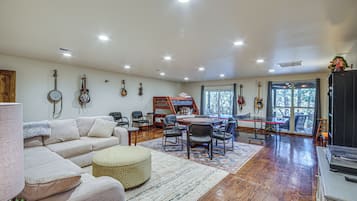 A spacious living room with a large sofa, a dining table, and several chairs. There are guitars hanging on the wall and a piano in the corner.