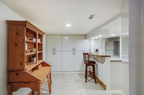 A compact kitchen with white cabinets, a wooden desk, and a small stool.