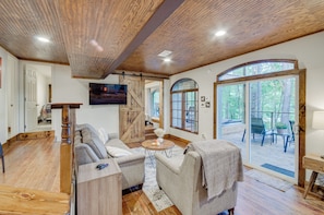A spacious living room with a wooden ceiling, a large window, and a sliding glass door leading to an outdoor deck. - Waterfront Home w/ 3 Decks, Fire Pits and Hot Tub! (Terry)