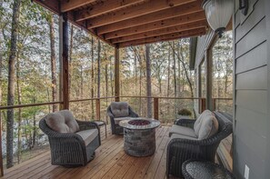 A covered deck with wicker furniture and a stone fire table, surrounded by tall trees.