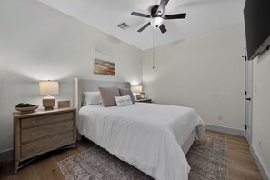 Un dormitorio con cama, mesitas de noche, ventilador de techo, un cuadro y una puerta.