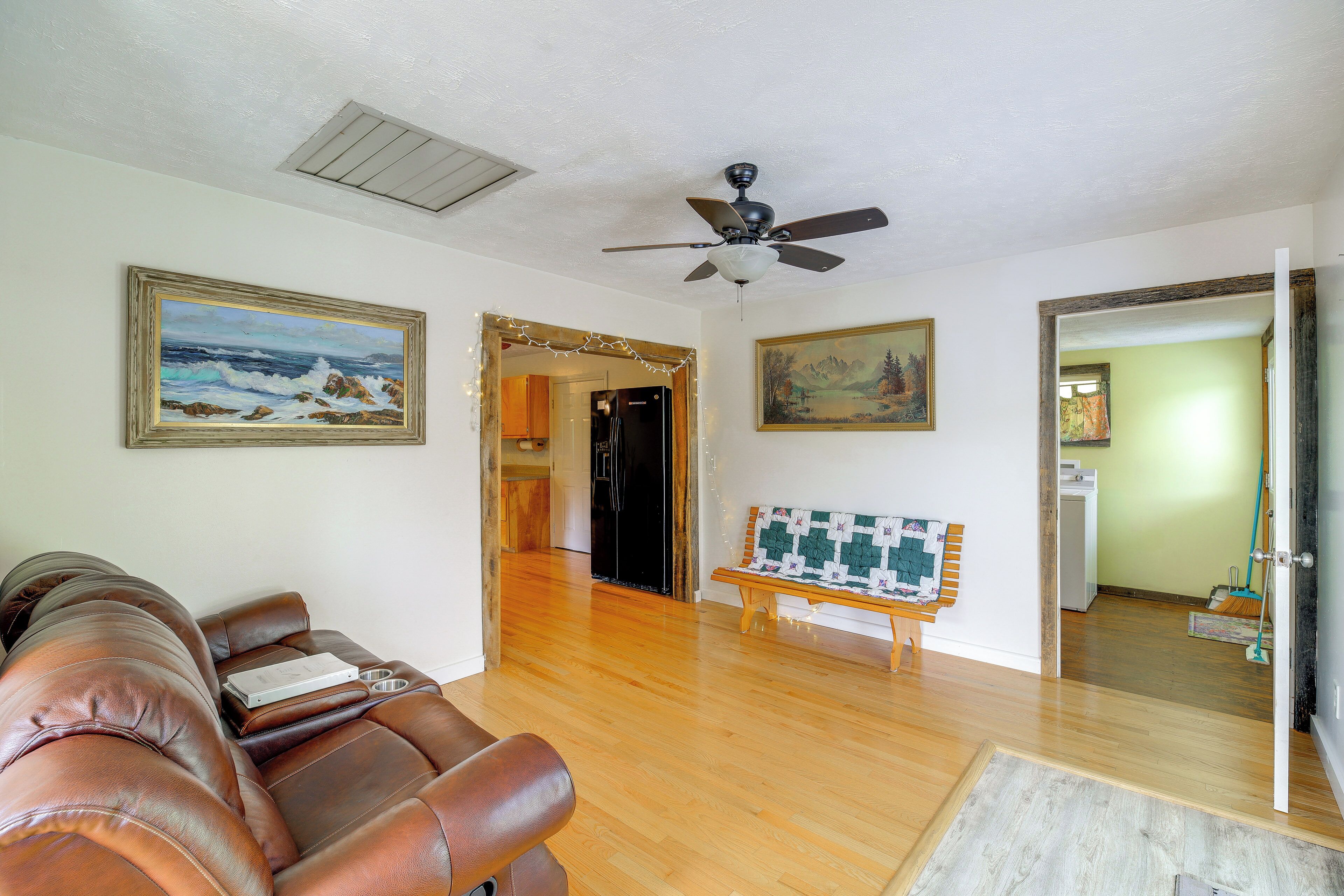 A living room with a brown leather sofa, wooden flooring, a ceiling fan, and two framed paintings.