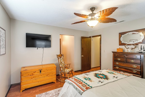 A bedroom with a ceiling fan, a bed with a patterned blanket, a wooden dresser, a chair, and a television on the wall.