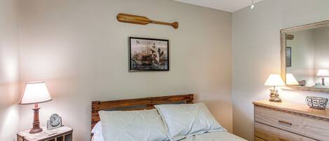 A bedroom with a wooden bed, a nightstand with a lamp, a framed picture on the wall, and a dresser with a mirror.