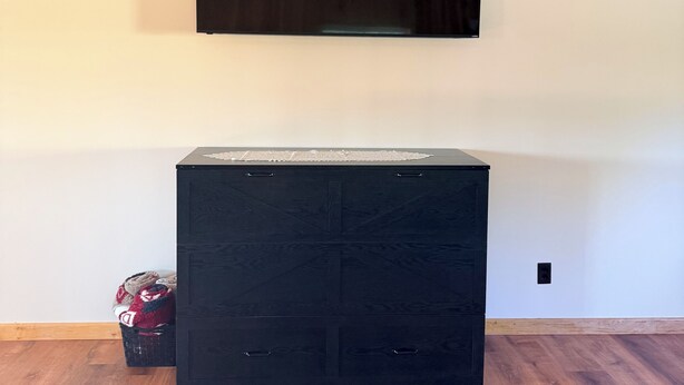 A room with a mounted television on the wall, a large black cabinet, and a wooden floor.