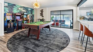 A modern living space with a pool table, arcade games, and a view to an outdoor patio. - Jungle Oasis - Pool + Hot Tub + Game Rooms (Richardson)
