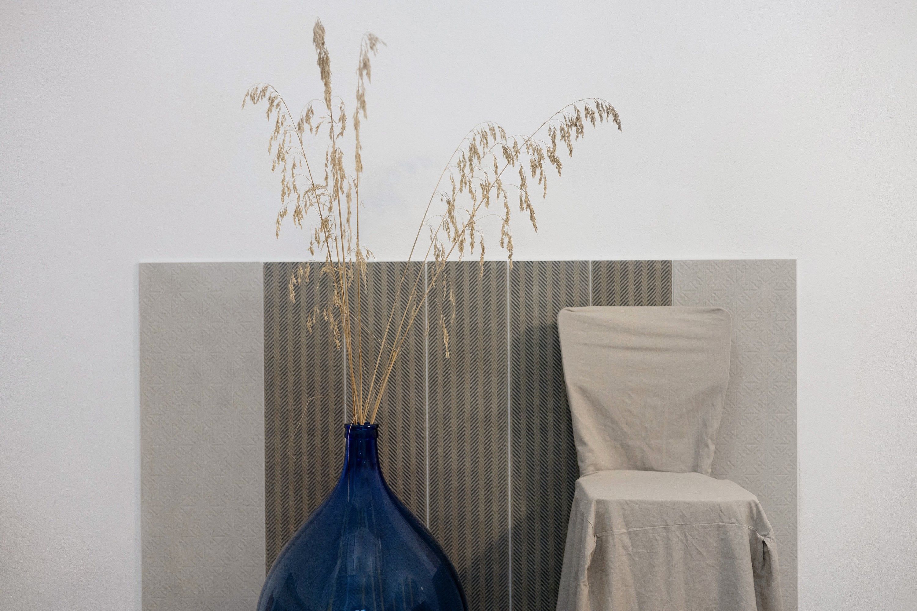 Ein Zimmer mit einer blauen Vase, in der getrocknete Pflanzen stehen, einer strukturierten Wand, einem schlichten Stuhl und einem gemusterten Wandteiler.
