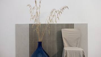 Ein Zimmer mit einer blauen Vase, in der getrocknete Pflanzen stehen, einer strukturierten Wand, einem schlichten Stuhl und einem gemusterten Wandteiler.