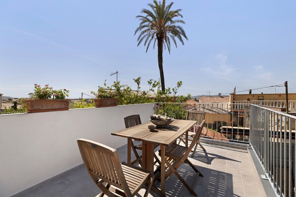 Un patio exterior con mesa y sillas de madera, rodeado de plantas en macetas y una palmera.