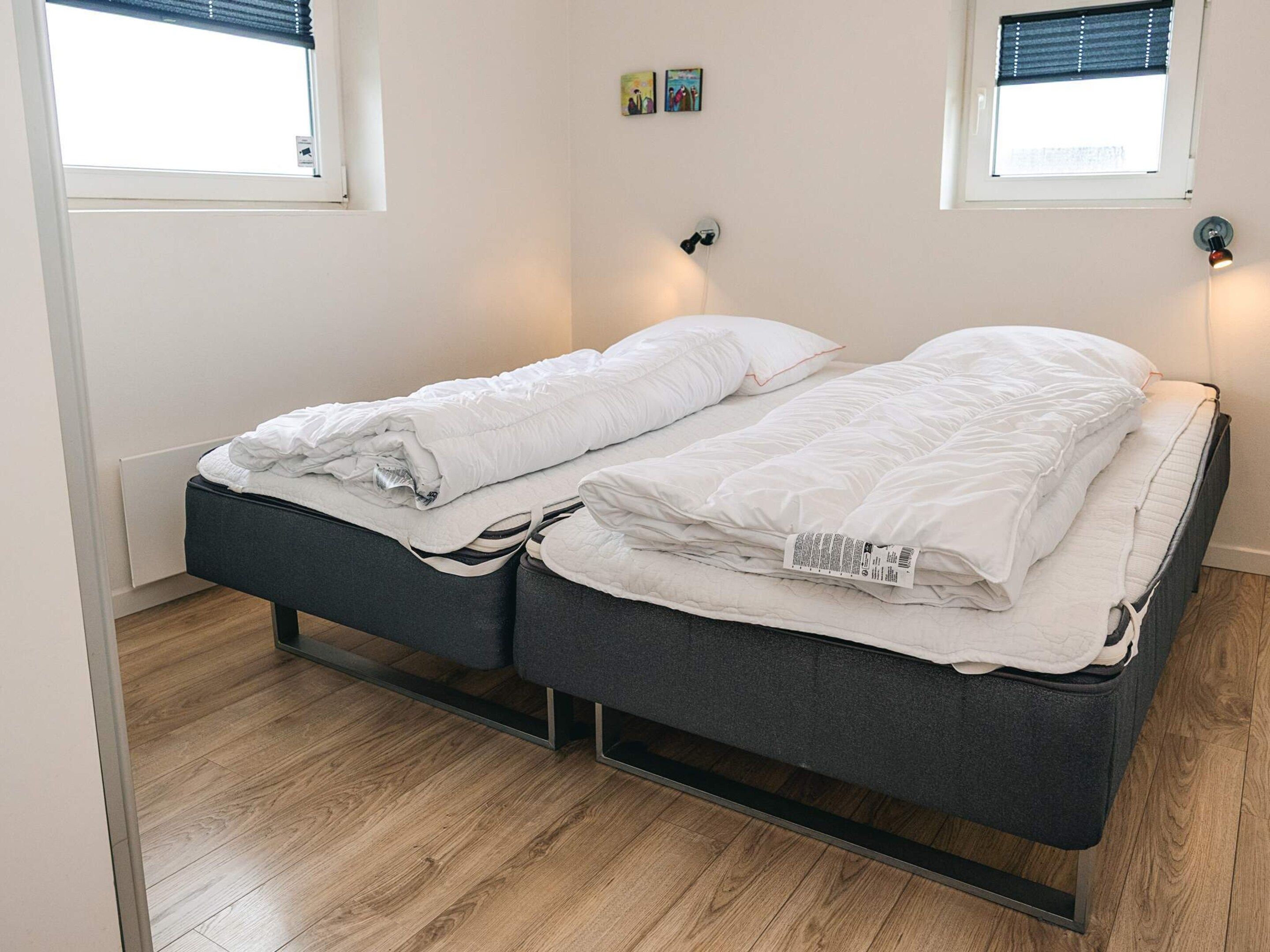 A single bed with a grey frame and white bedding, two wall-mounted lights, and a window with blinds.