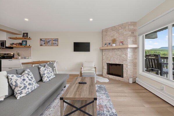 A modern living room with a fireplace, a sofa, a coffee table, and a large window offering a view of the outdoors. - Winter Park Lodge II Unit E102 Renovated, mt views, 2 bedroom with amenities (Fraser)