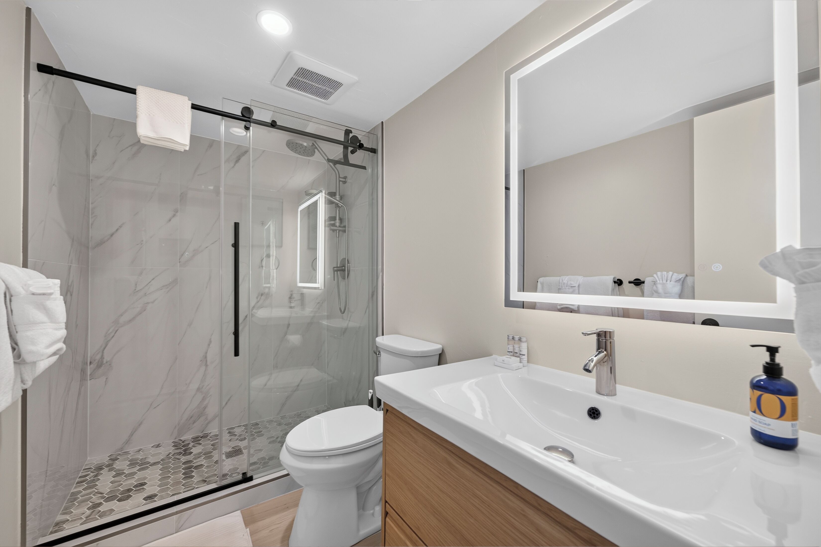 A modern bathroom with a glass-enclosed shower, a white toilet, a large sink with a mirror, and a bottle of soap.