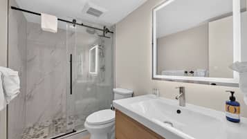 A modern bathroom with a glass-enclosed shower, a white toilet, a large sink with a mirror, and a bottle of soap.
