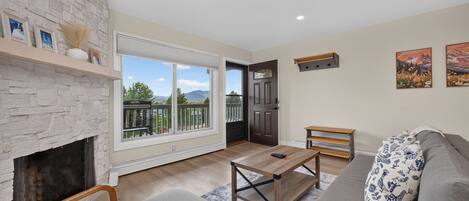 A living room with a stone fireplace, a sofa, a coffee table, and a view of a mountain landscape through large windows.