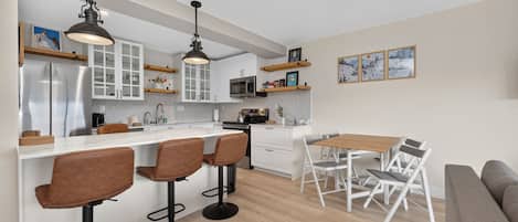 Eine moderne Küche mit weißen Schränken, einer zentralen Kochinsel mit Barhocker, einem Essbereich mit Tisch und Stühlen sowie einem Wohnbereich mit einer Couch.