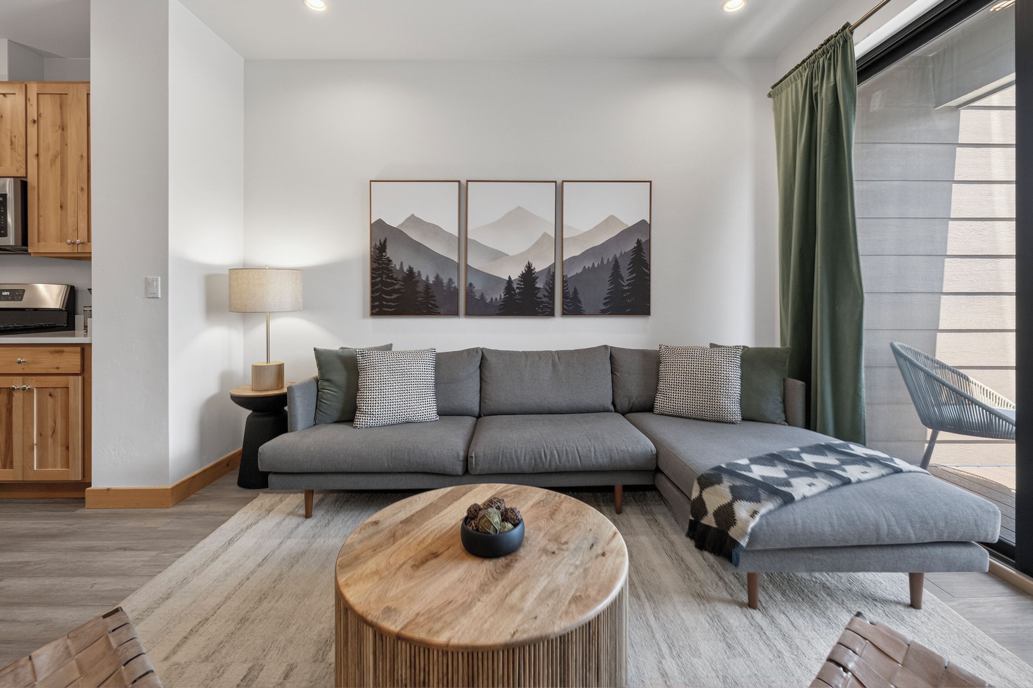 A modern living room with a grey sectional sofa, a round wooden coffee table, and a mountain landscape painting above the sofa.