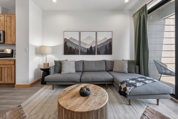 Ein modernes Wohnzimmer mit einem grauen Ecksofa, einem runden Holztisch und einem Gemälde von einer Berglandschaft darüber.