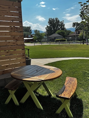 A wooden outdoor seating area with a table and bench, a grassy lawn, and houses in the background.