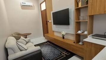 A modern living room with a sofa, a coffee table, a flat-screen TV mounted on a wooden wall, a small shelf with cups, and a decorative vase.
