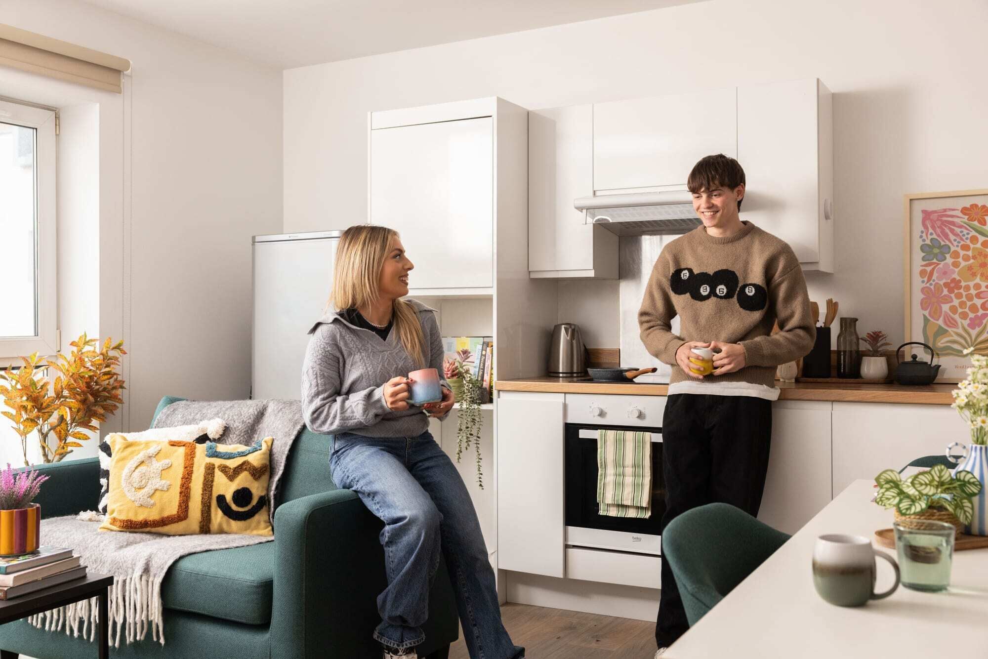 A modern kitchen with white cabinets, a stove, and a countertop. There is a person standing near the stove, and another person sitting on a couch with a decorative pillow.
