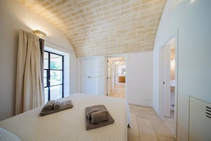 A bedroom with a large bed, a window with curtains, a hallway leading to a bathroom, and a brick ceiling. - TD Villa Antonia - Hidden Citrus Retreat (Ostuni)