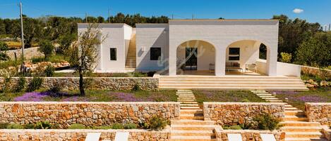 Un bâtiment moderne avec une façade en pierre, une entrée centrale avec des marches et un jardin agrémenté de fleurs violettes.