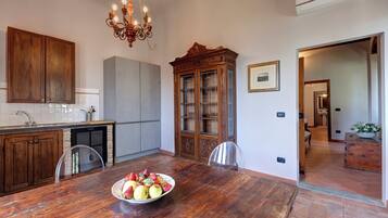 A dining area with a wooden table, chairs, and a bowl of fruit. There is a chandelier, cabinets, and an air conditioning unit.