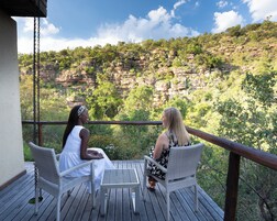 Two people seated on wicker chairs on a wooden deck with a table, overlooking a rocky cliff with vegetation.