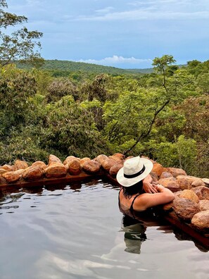 A person sitting on rocks by a calm body of water, surrounded by lush greenery and hills in the distance. - The Riverine Lodge  (Vaalwater)