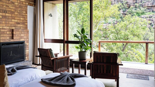 Una habitación con chimenea, dos sillas de madera, una mesita, una planta en maceta y una cama con manta.