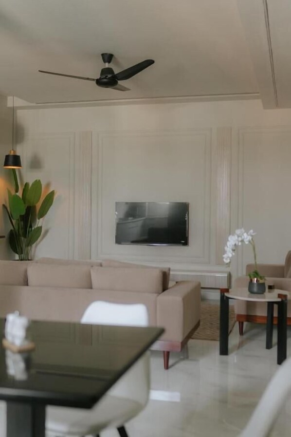 Een woonkamer met een beige bank, een aan de muur bevestigde televisie, een plafondventilator, een eettafel met stoelen en een potplant.