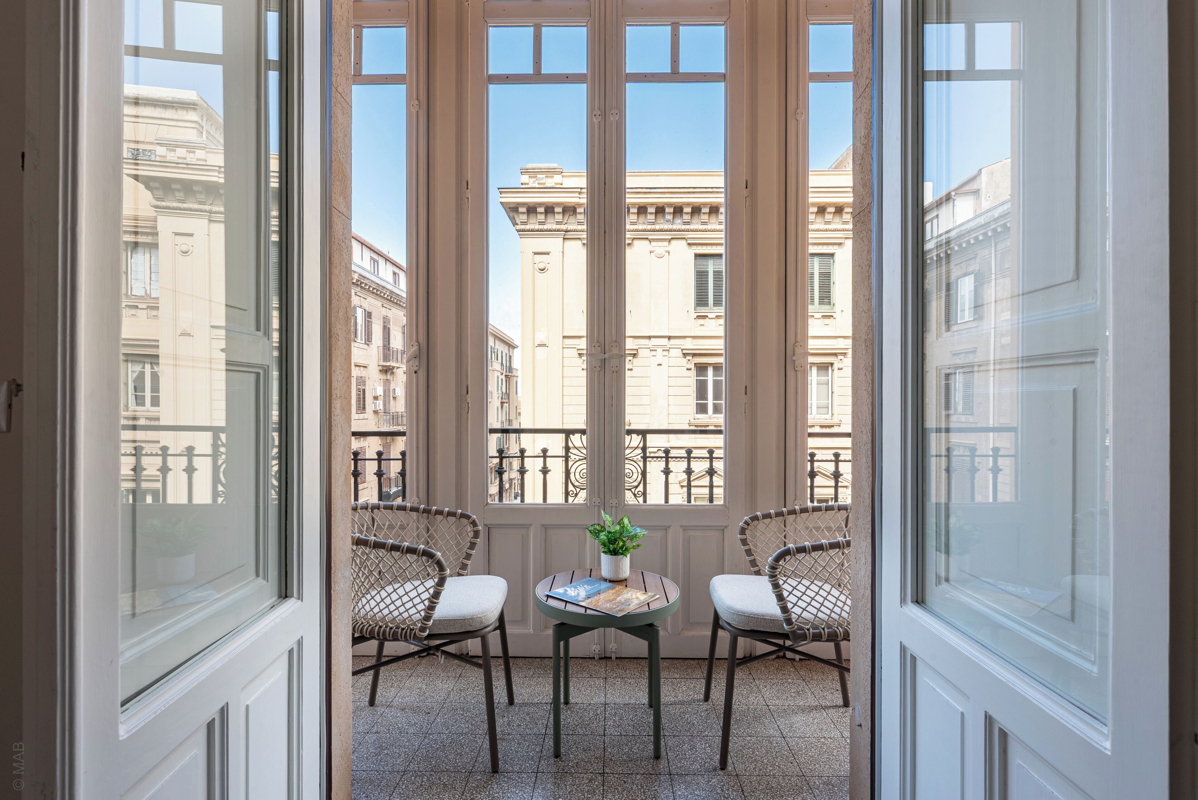 Een balkon met twee rieten stoelen, een tafeltje met een plant, en uitzicht op klassieke gebouwen.