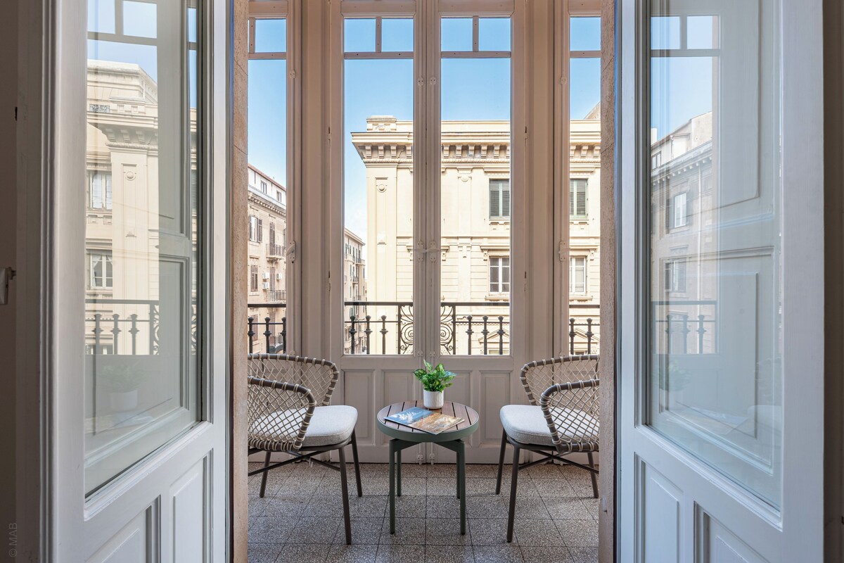 A balcony with two wicker chairs, a small table with a plant, and a view of classical buildings.