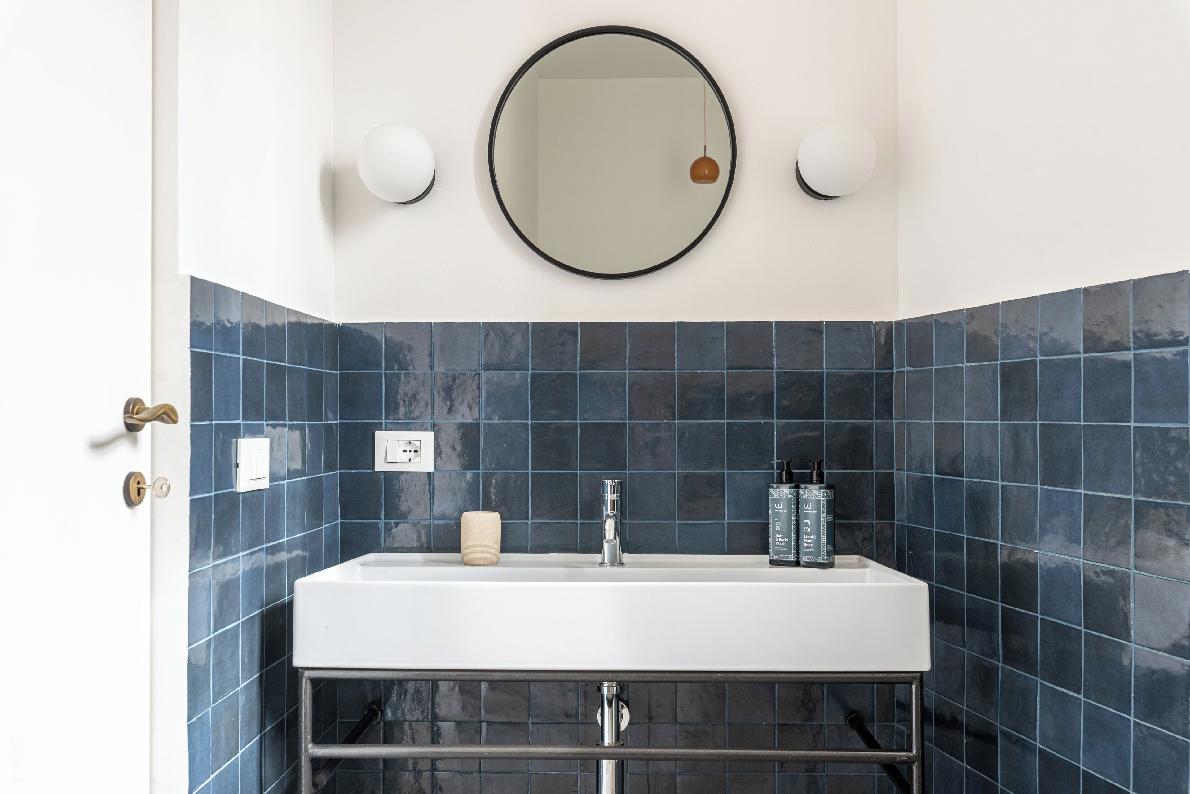 A bathroom with a white sink, dark blue tiles, a round mirror, and two wall-mounted lights.