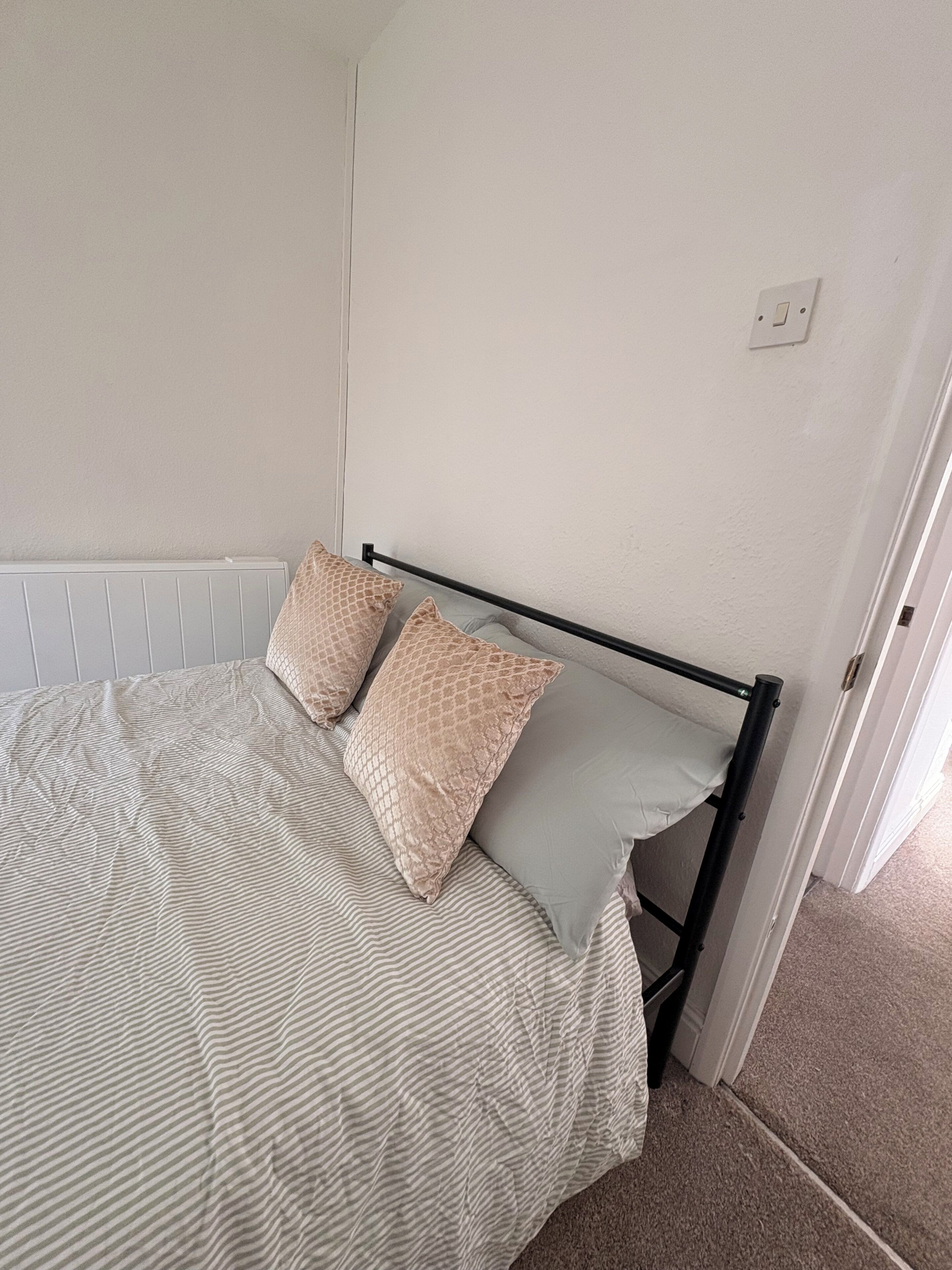 A bed with a striped bedspread and two patterned pillows, a radiator, and a light switch on the wall.