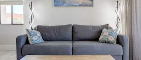 A living room with a grey sofa, two patterned pillows, a wooden coffee table, and a painting of a beach scene on the wall.