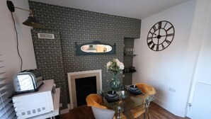 Un comedor con una mesa de vidrio, dos sillas, un jarrón con flores, un reloj y una chimenea.