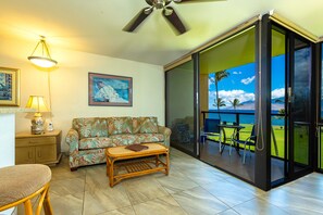 A living room with a sofa, coffee table, and a view of a tropical landscape through a sliding glass door. - Kihei Surfside #311 1 Bedroom Condo (Kihei)